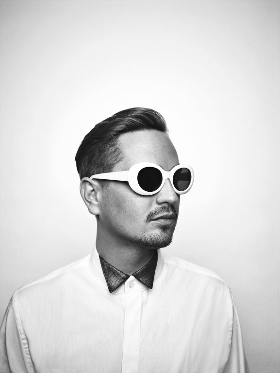 A black and white image of chart hit artist Robin Schulz. He is wearing a shirt and large, white sunglasses. He looks to the right with a neutral expression